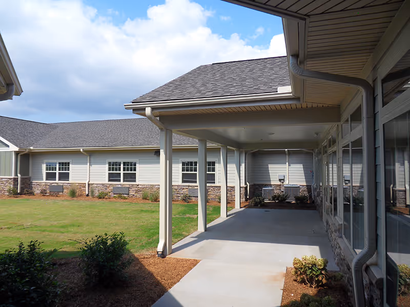 Covered walkway outside a single-story building with beige siding and stone accents, surrounded by a grassy area and small shrubs under a partly cloudy sky.