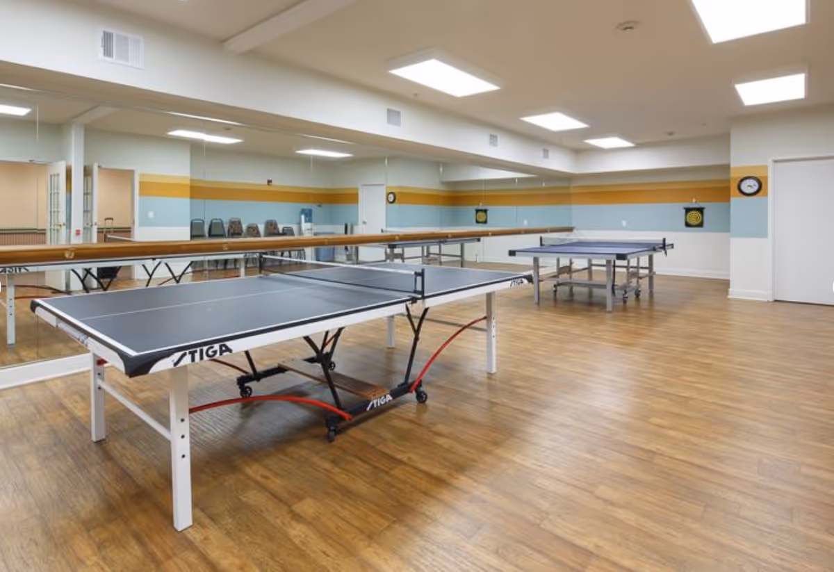 A spacious indoor recreational room with two ping pong tables on a wooden floor. The walls have a horizontal stripe design in yellow and light blue, and one wall is covered with large mirrors. There are several chairs lined up against the wall and a clock and dartboard mounted on the far wall.