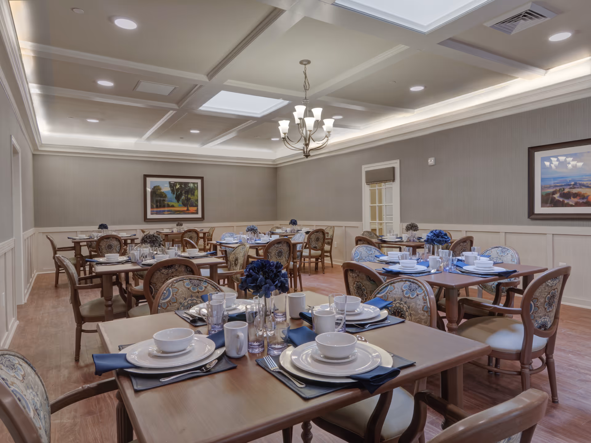 A well-lit dining room with multiple wooden tables set with white plates, bowls, cups, and silverware on dark placemats. Each table has a centerpiece of blue flowers. The room features upholstered chairs with patterned fabric, a chandelier hanging from a coffered ceiling with recessed lighting, and framed landscape paintings on the walls.