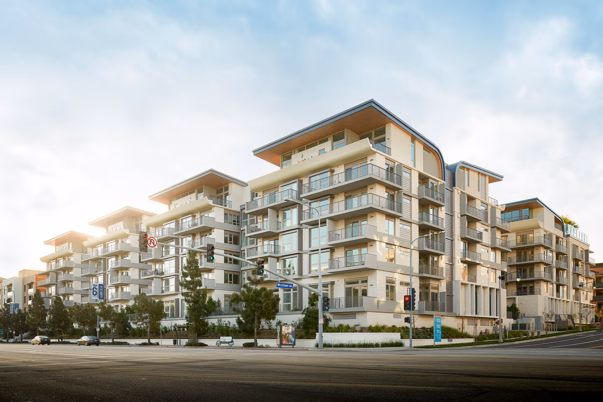 Modern multi-story residential building with balconies, large windows, and a clean architectural design, situated at a street intersection with traffic lights and some trees along the sidewalk under a partly cloudy sky.