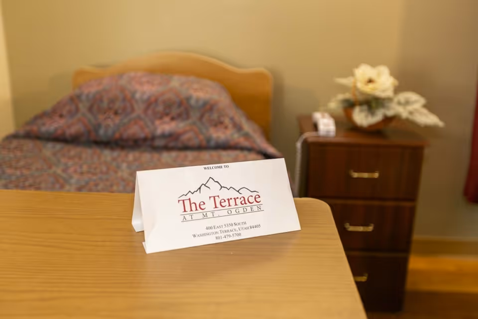 A tabletop welcome card for The Terrace at Mt. Ogden sits on a table in a bedroom with a bed and nightstand.