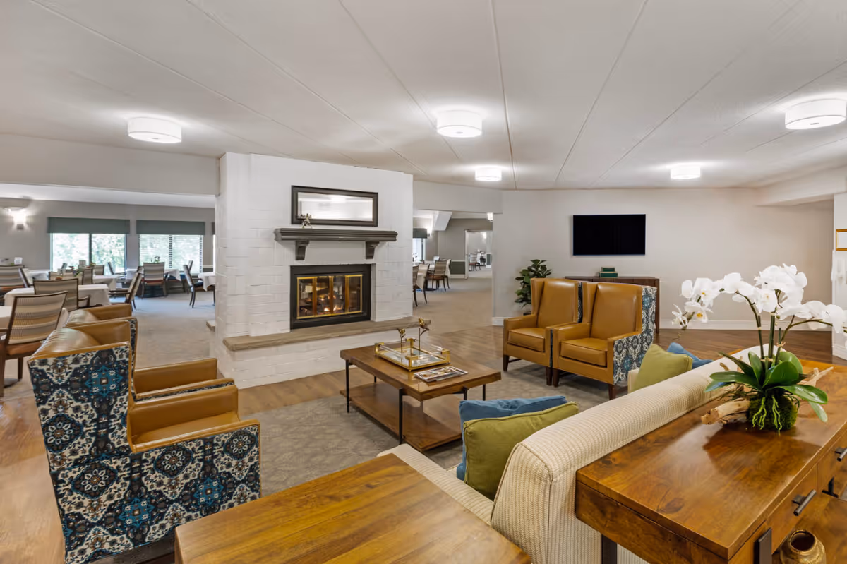 A cozy senior living common area featuring a white brick fireplace with a mirror above it, surrounded by comfortable seating including patterned armchairs and tan leather chairs. A wooden coffee table with decorative items sits in the center on a rug. In the background, there is a dining area with tables and chairs near large windows, and a flat-screen TV mounted on the wall. The room has a warm, inviting atmosphere with wood flooring and soft lighting.