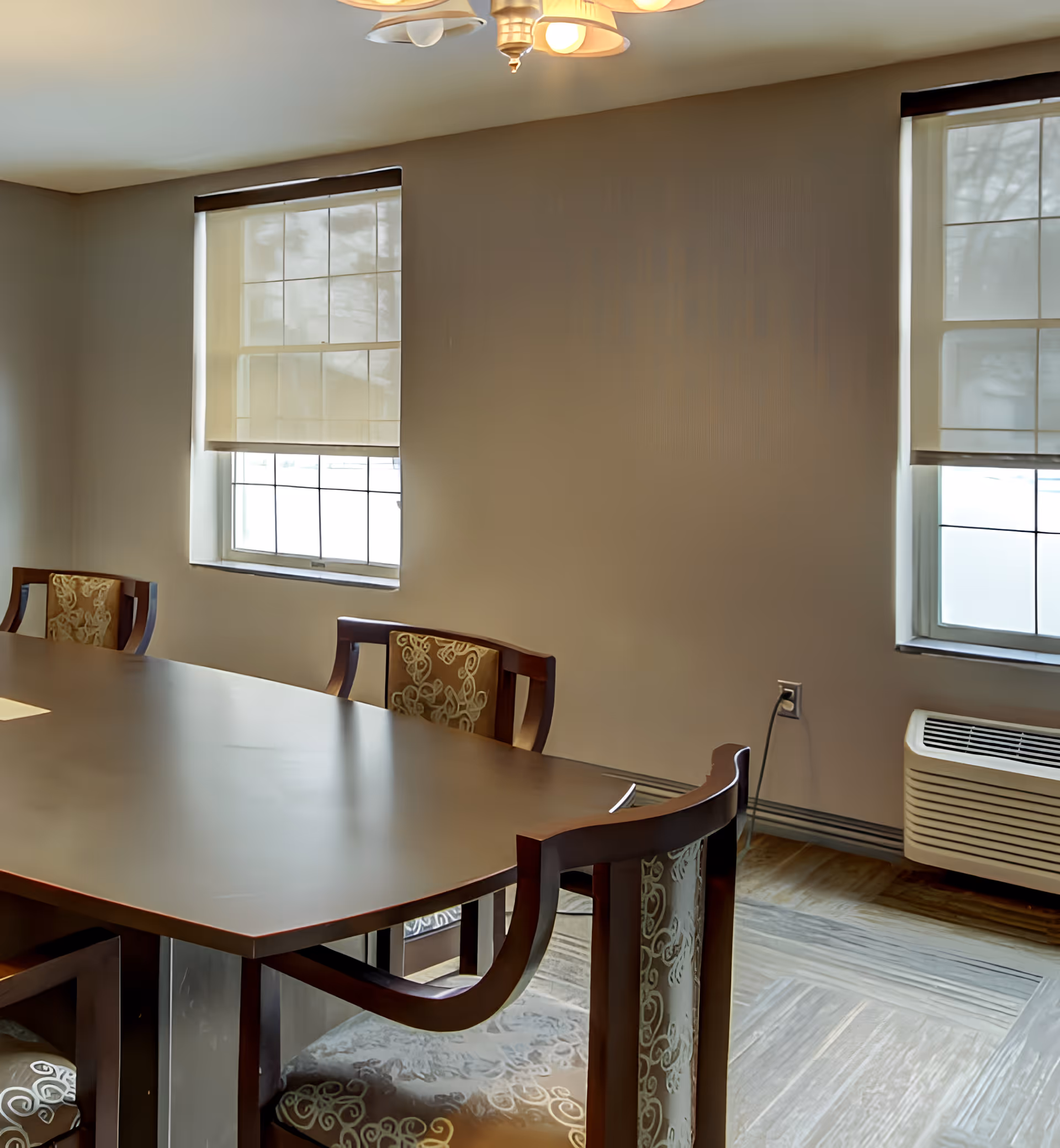A wooden dining table surrounded by upholstered chairs in a bright room with two windows and a wall air conditioning unit.