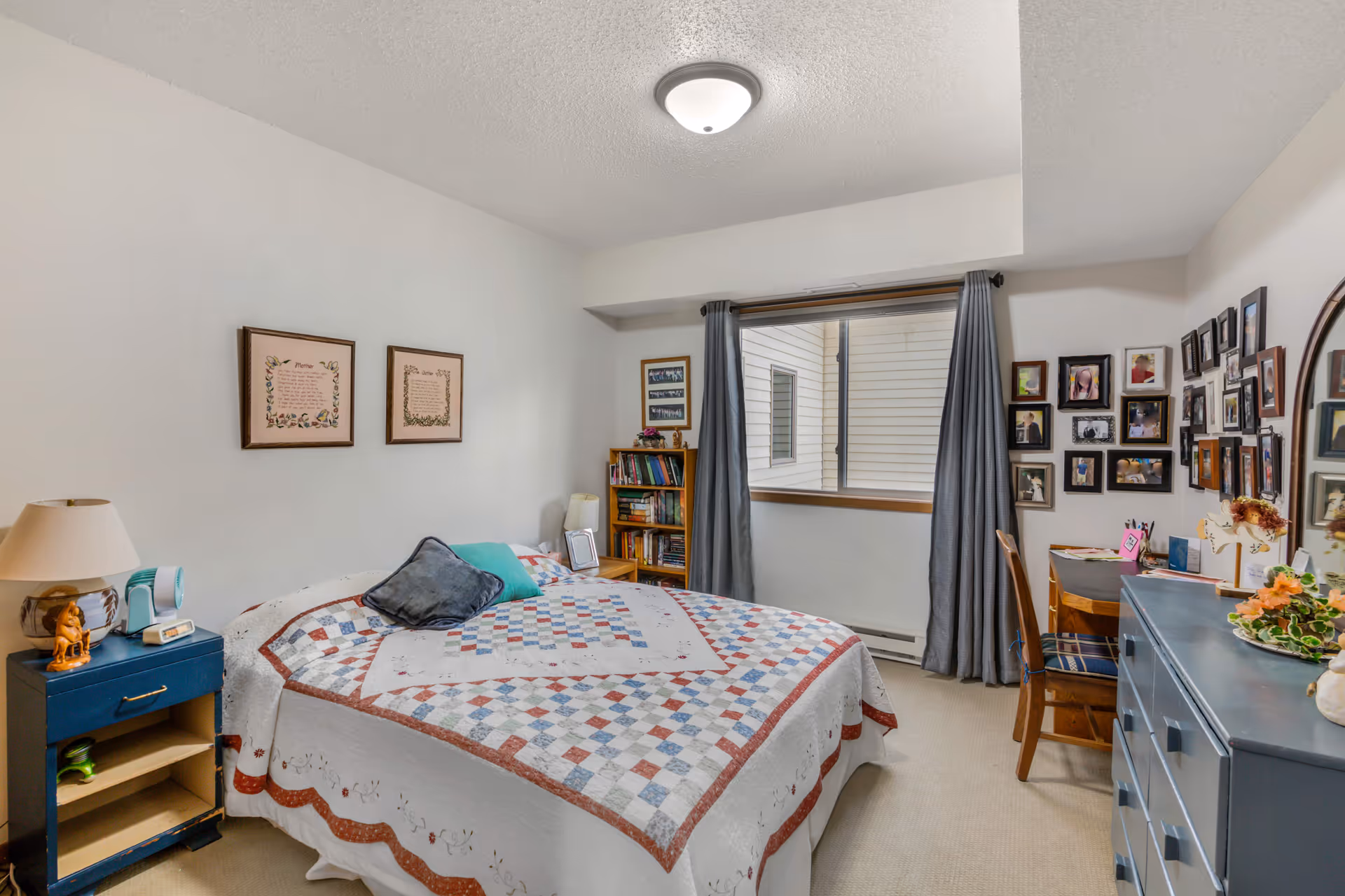 Furnished bedroom with a quilt-covered bed, nightstands, dresser, window with curtains, bookshelf and a wall of framed photos.