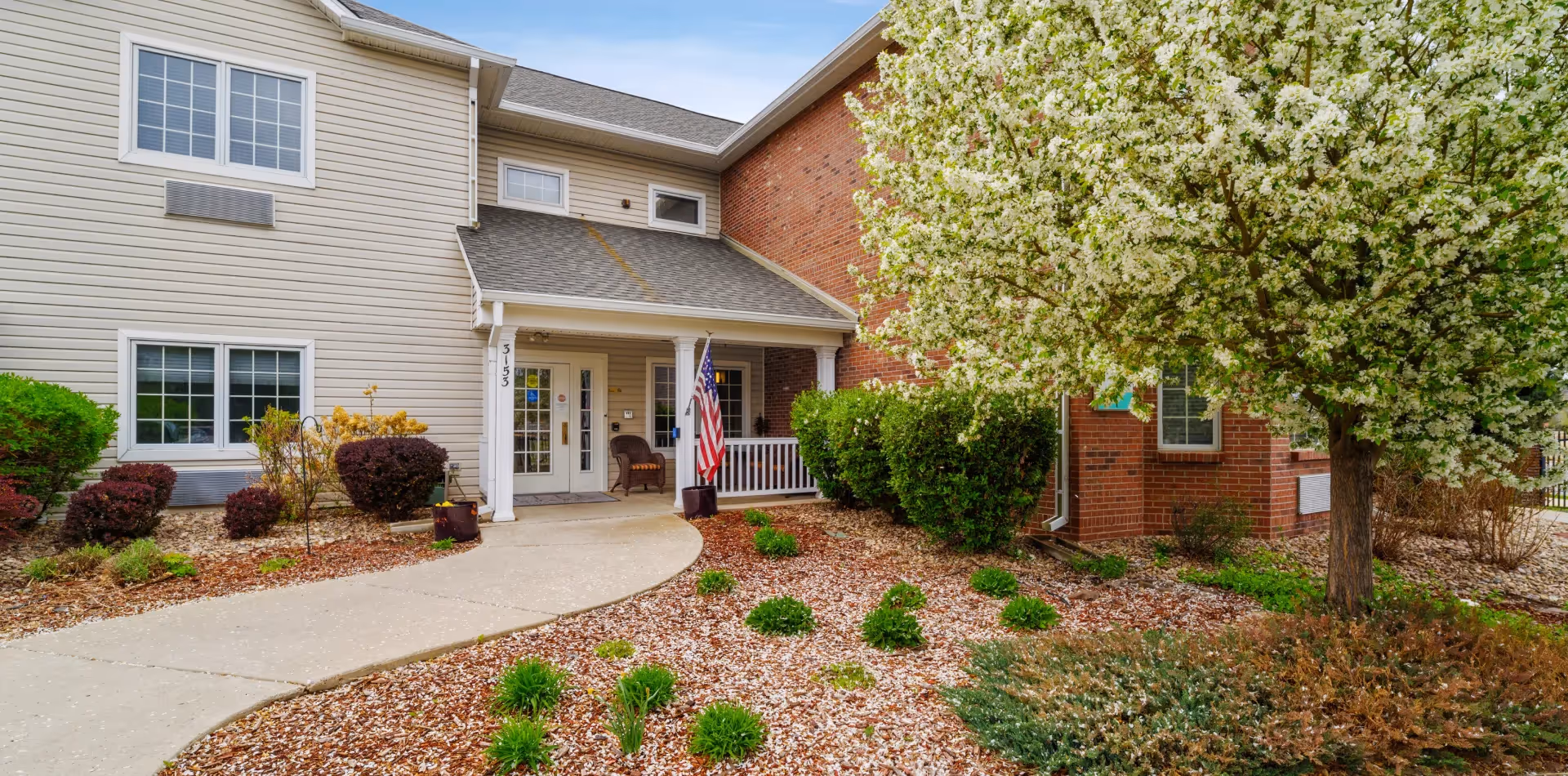 Front entrance of a senior living building with a covered porch, American flag, landscaped beds and a blooming tree.