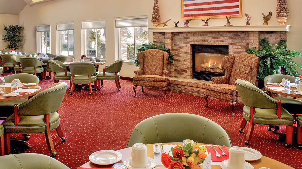 A dining room with multiple round tables set with plates, cups, and silverware. The room has green cushioned chairs on wheels, a red carpet with a subtle pattern, and a brick fireplace with a fire burning. Above the fireplace is an American flag and decorative eagle figurines. Large windows let in natural light and there are green plants near the fireplace.
