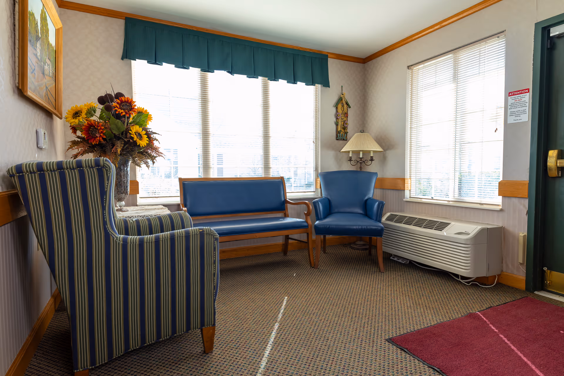 A small sitting area with a striped armchair, a blue cushioned bench, and a blue armchair arranged near two windows with blinds. A vase with sunflowers and other flowers is on a small table next to the armchair. A floor lamp is placed in the corner, and a framed painting hangs on the wall. The room has carpeted flooring and a maroon rug near a green door.