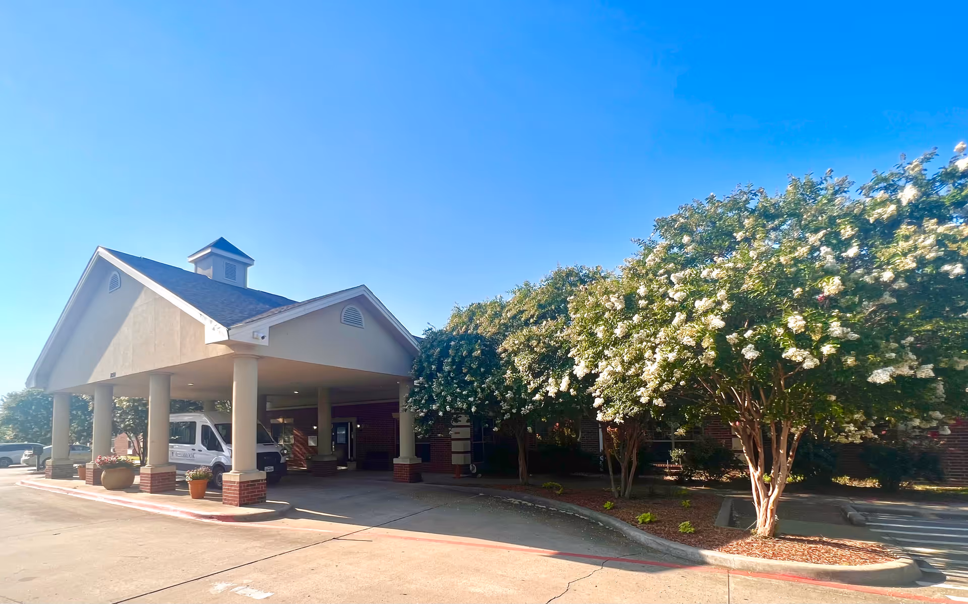Exterior view of Millbrook Healthcare and Rehabilitation Center showing the entrance with a covered drop-off area supported by columns. There are flowering trees and landscaping along the driveway under a clear blue sky.