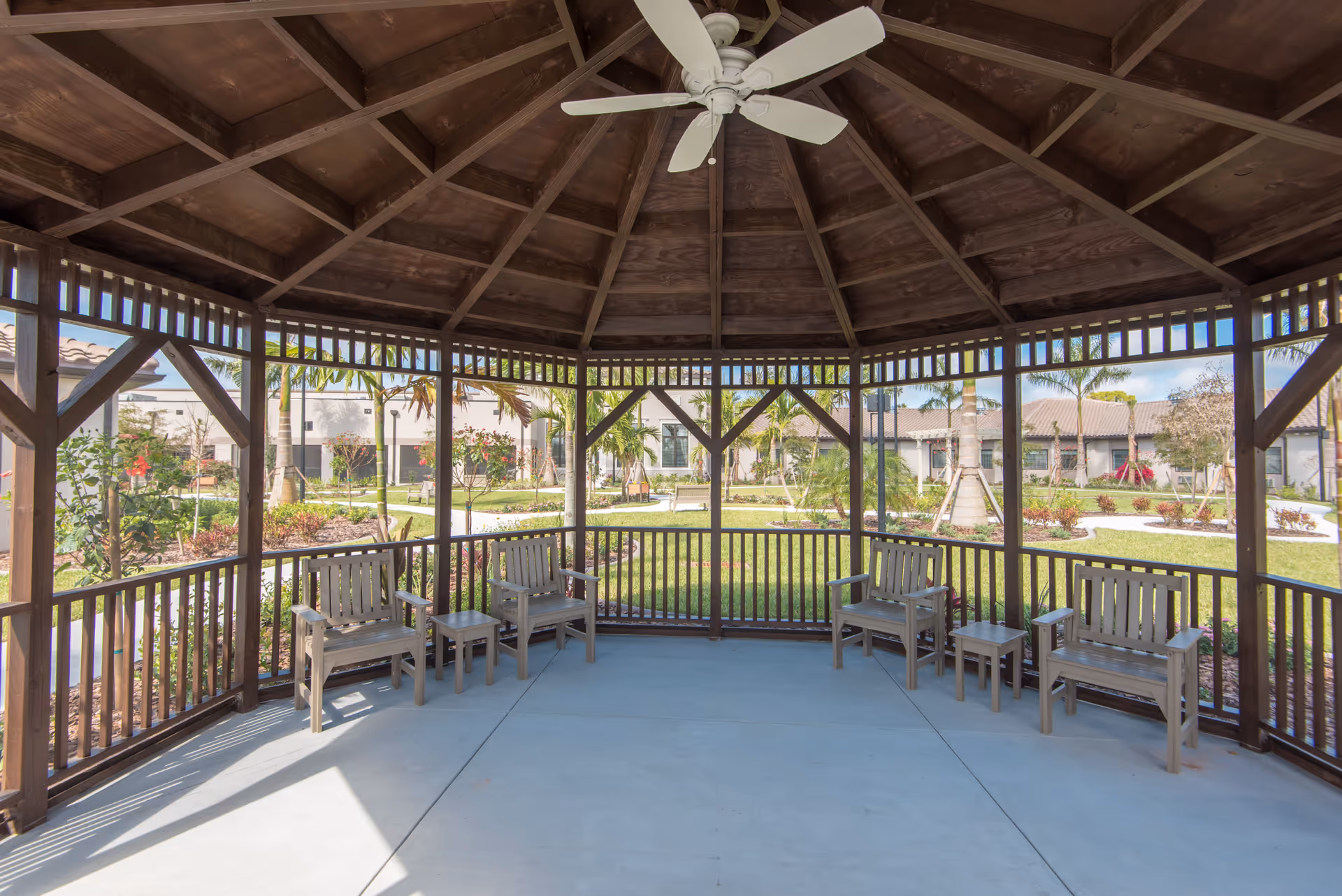 View from inside a wooden gazebo with a ceiling fan, featuring several wooden benches and small tables. The gazebo overlooks a landscaped garden area with palm trees, shrubs, and a pathway, with buildings visible in the background under a clear sky.