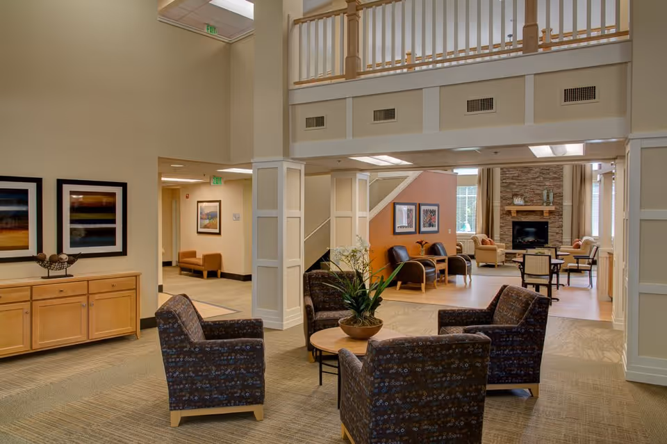 A spacious and well-lit assisted living facility common area with four patterned armchairs arranged around a round wooden table with a potted plant. The room features beige walls, carpeted flooring, and a wooden railing on the upper level. In the background, there is a sitting area with additional chairs, a stone fireplace, and framed artwork on the walls.