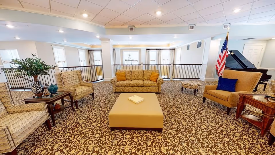 Spacious communal lounge with patterned carpet, sofas, armchairs, an ottoman, piano and an American flag.