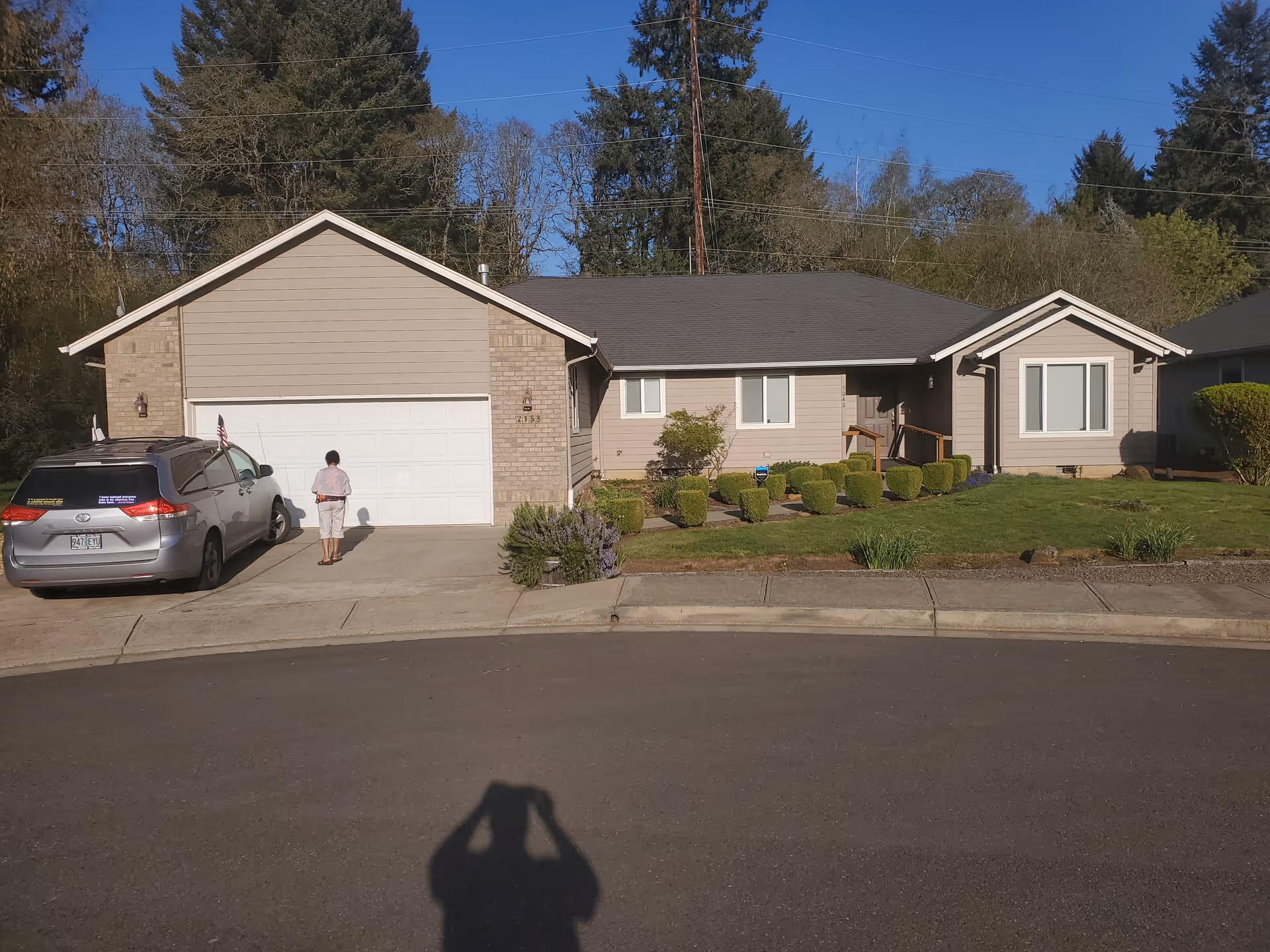 Single-story residential house with beige siding and brick accents, a two-car garage, a silver minivan parked in the driveway, and a person standing near the garage door. The front yard has neatly trimmed bushes and a green lawn, with tall trees in the background under a clear blue sky.
