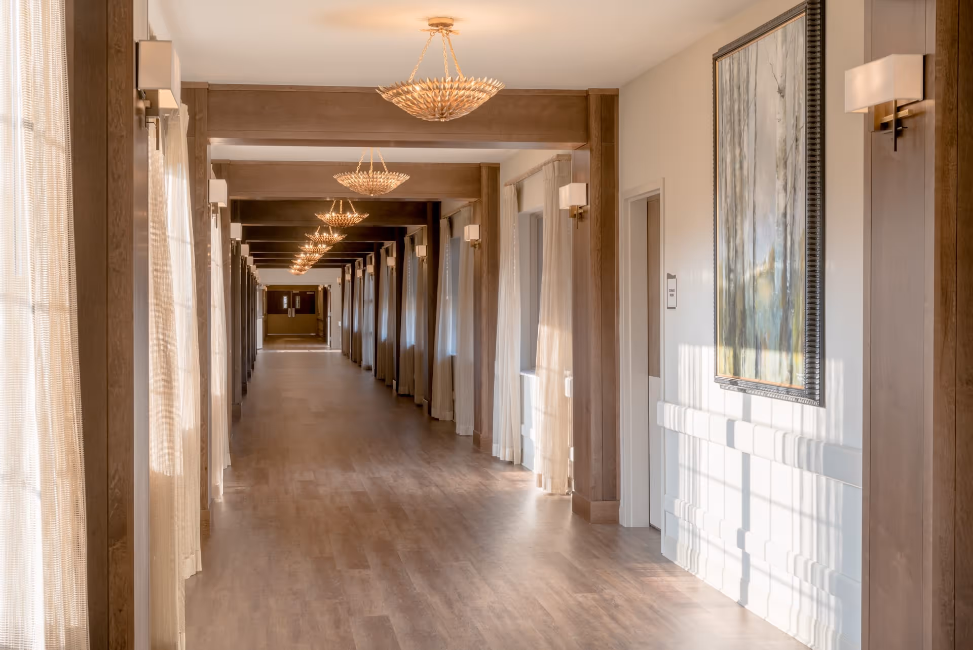 A long, well-lit hallway with wooden floors and beams, large windows with sheer curtains on the left side, and wall sconces and chandeliers providing warm lighting. A large framed painting is hung on the right wall near a door.