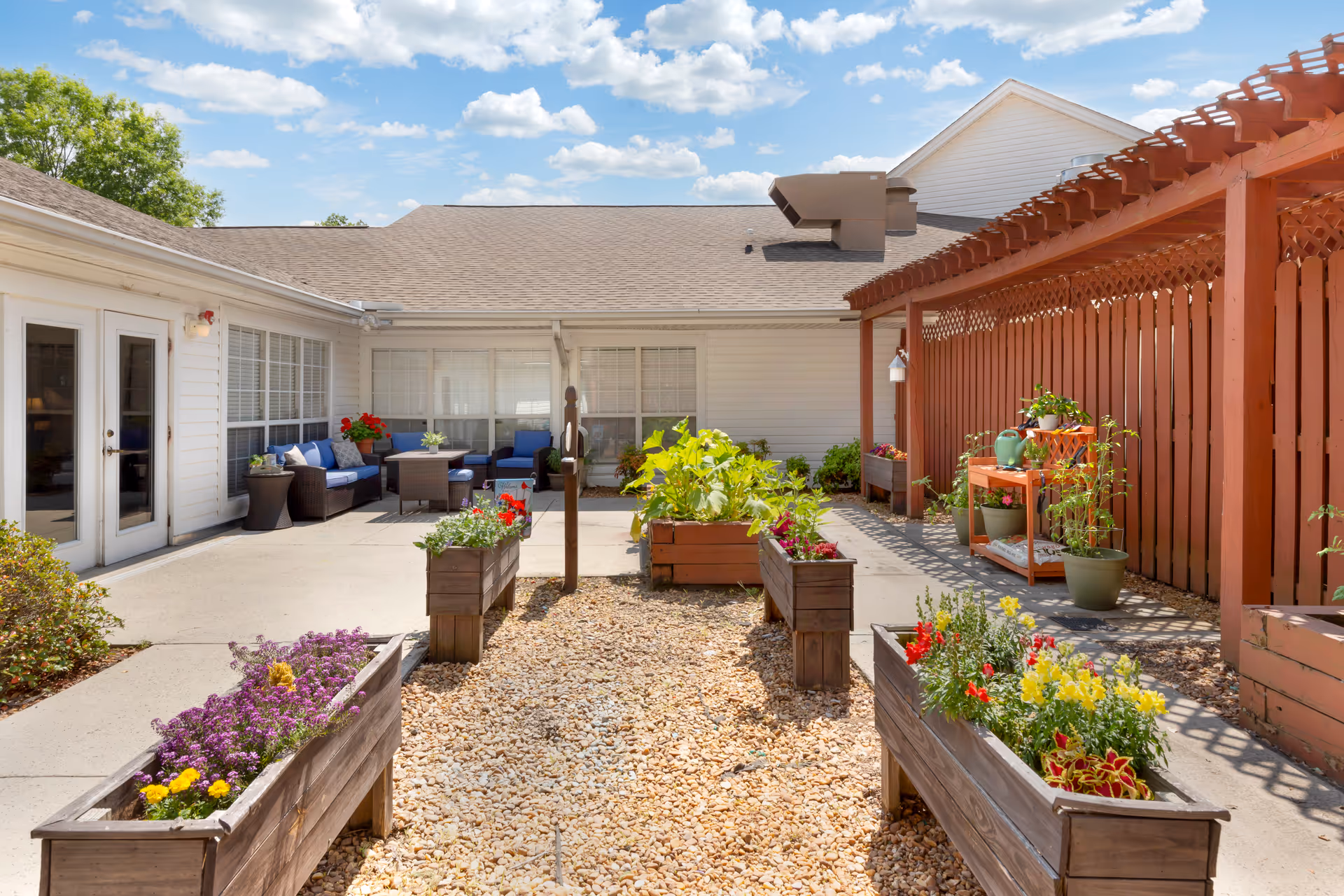 Sunny courtyard with raised wooden planters, potted plants, a seating area and a pergola beside a single-story building.