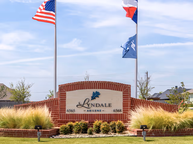 Brick entrance monument sign reading "Lyndale Abilene" surrounded by landscaping and flagpoles under a blue sky.