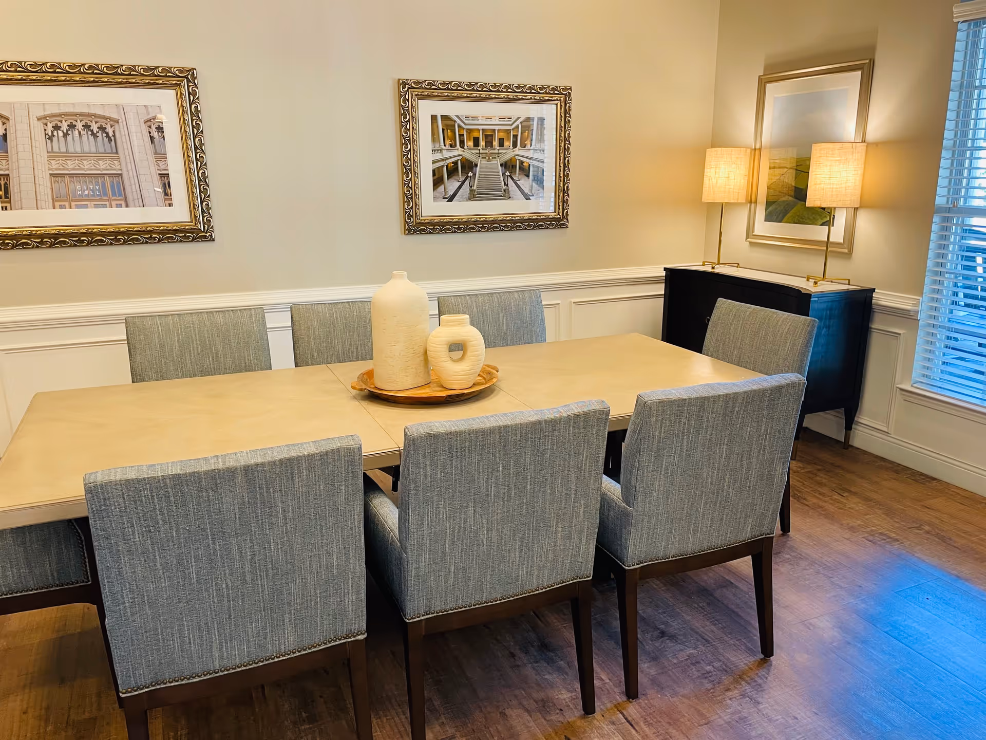 A dining room with a rectangular table surrounded by eight upholstered chairs. On the table are two decorative vases on a wooden tray. The room has wooden flooring, two framed pictures on the wall, a sideboard with two lamps, and a window with blinds letting in natural light.