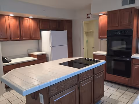 A spacious kitchen with a tiled island cooktop, wooden cabinets, a white refrigerator and a double oven.