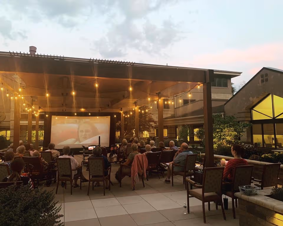 A group of senior residents seated outdoors under a pergola with string lights, watching a movie projected on a large screen during dusk at Harwood Place Senior Living.