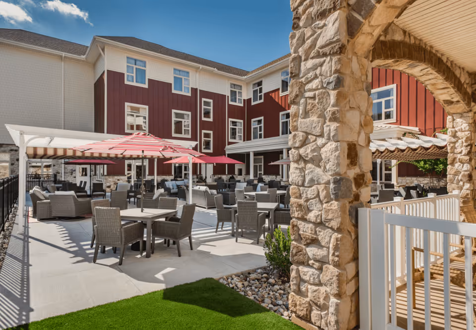 Outdoor patio area at Fieldstone at Chester Springs featuring multiple tables with chairs and umbrellas, surrounded by a three-story building with red and white siding. Stone pillars and a white railing are visible in the foreground under a clear blue sky.
