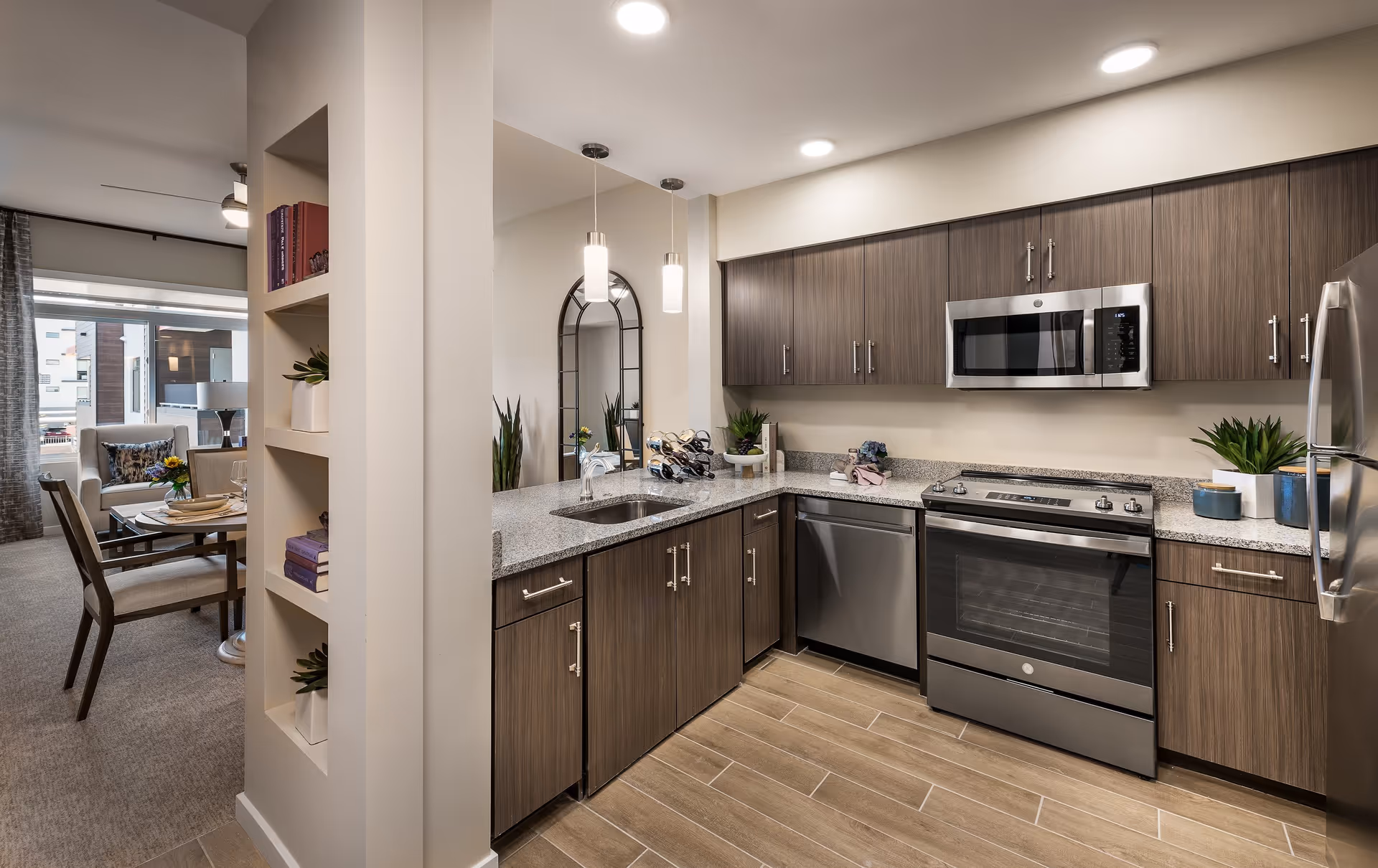Modern kitchen with wood cabinets, granite countertops, stainless steel appliances including a microwave, dishwasher, and oven. The kitchen opens to a dining area with a round table and chairs, and a living area with a large window and armchair. Decorative plants and books are placed on shelves and countertops.