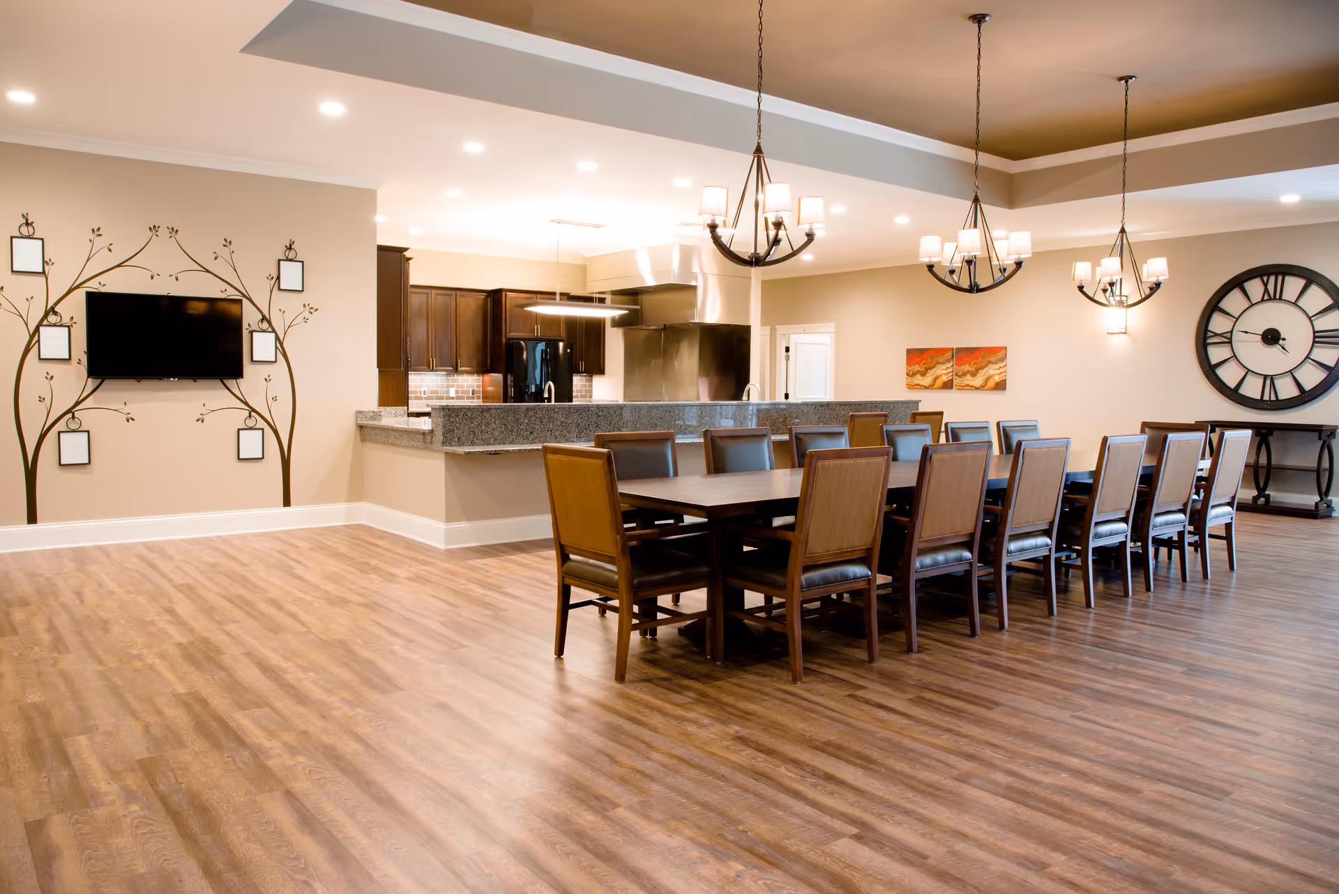 A spacious dining room with a long wooden table surrounded by twelve chairs with dark cushions. The room features wood flooring, three hanging chandeliers, a large wall clock, and a kitchen area with granite countertops and dark wood cabinets in the background. There is also a wall-mounted TV with decorative frames arranged around it on one wall.