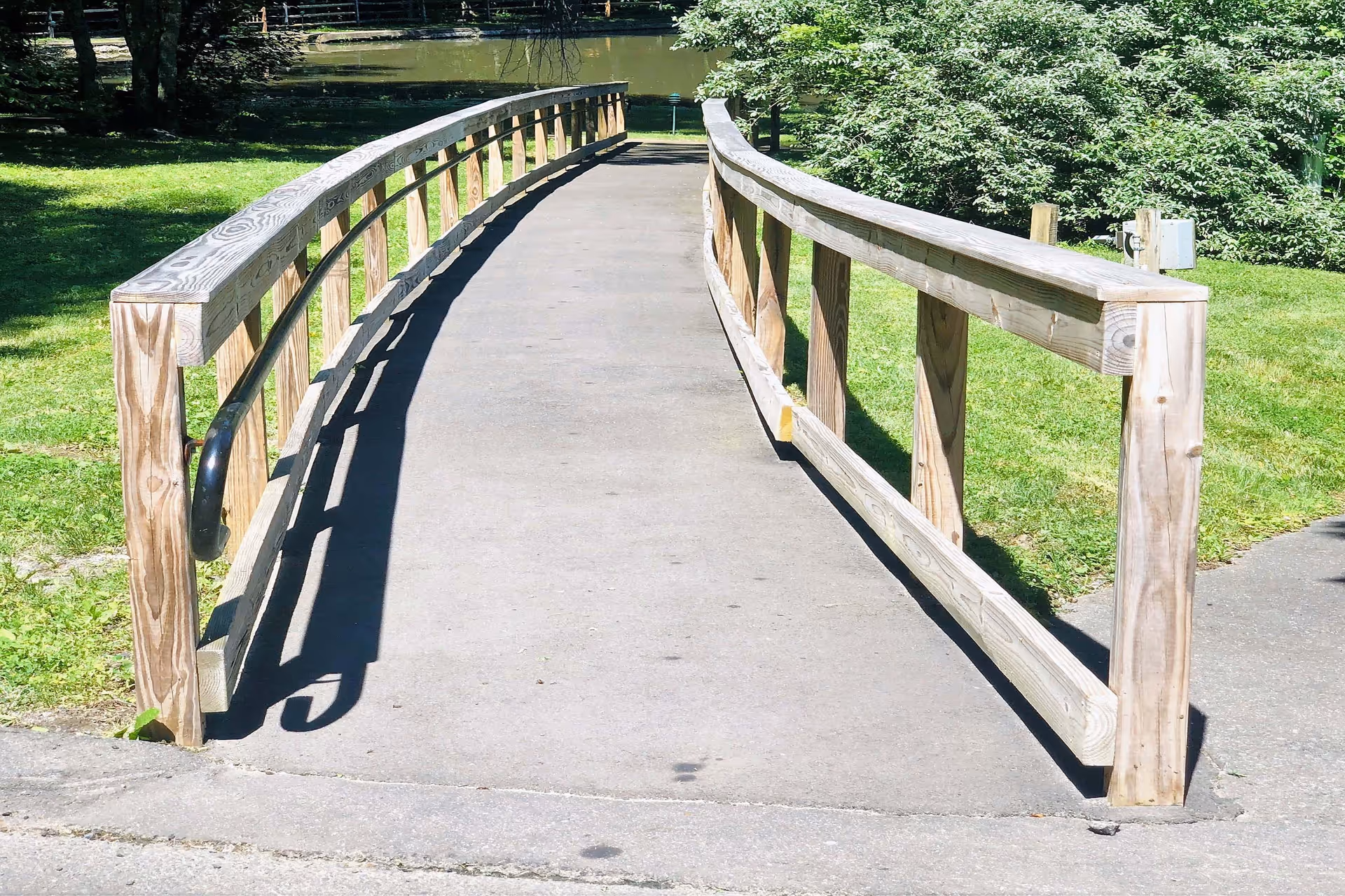 Wooden pedestrian bridge with railings crossing a paved path in a grassy park.
