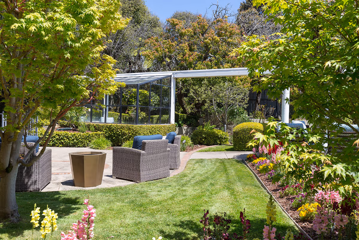 Sunlit outdoor garden patio with wicker chairs, a small table, manicured lawn, and colorful flowerbeds.