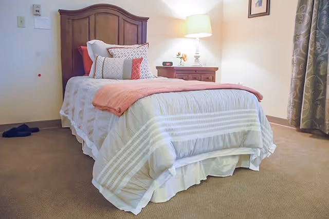A neatly made single bed with a wooden headboard in a carpeted room. The bed has white and light gray bedding with a coral throw blanket and several decorative pillows. Next to the bed is a wooden nightstand with a lamp, a small clock, and a flower vase. A curtain hangs on the right side of the image, and a framed picture is on the wall above the nightstand.