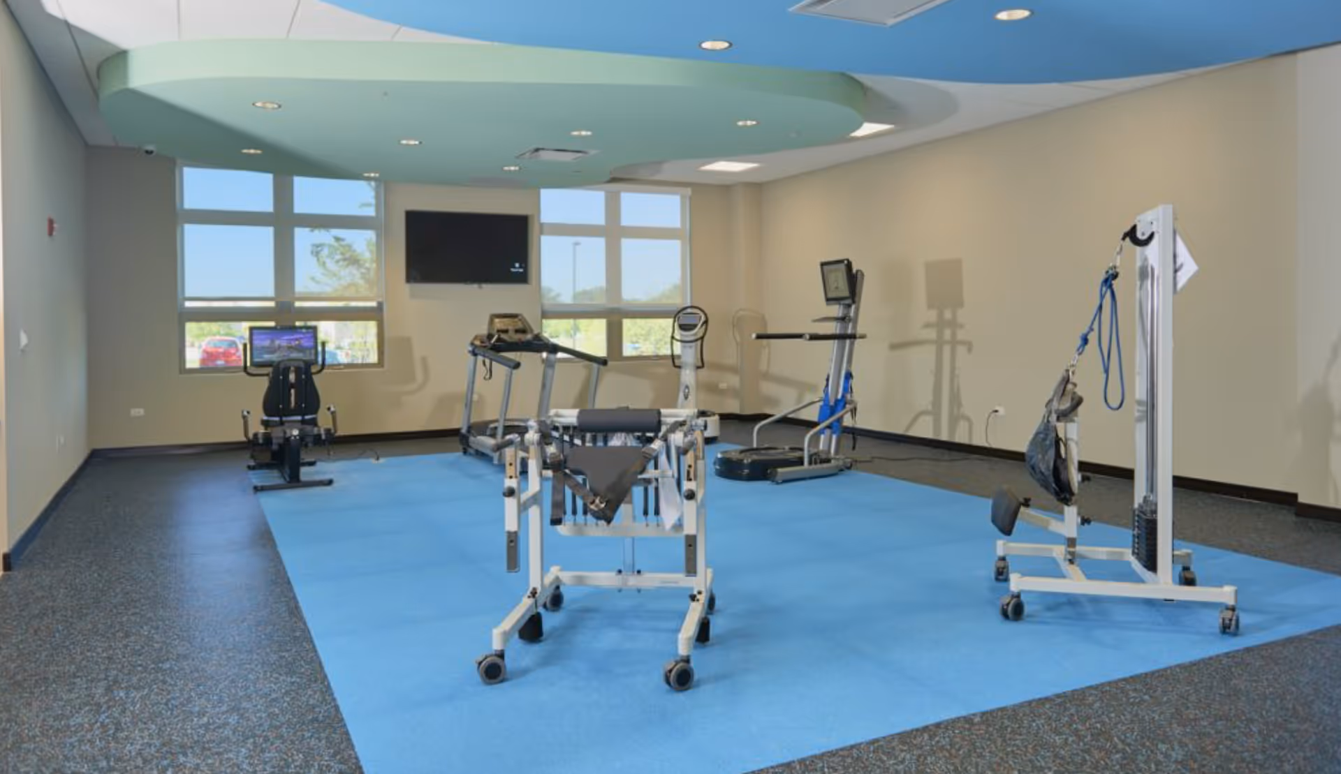 A rehabilitation or physical therapy room with exercise equipment including a recumbent bike, treadmill, and various assistive devices on a blue mat. The room has large windows letting in natural light and a wall-mounted TV.