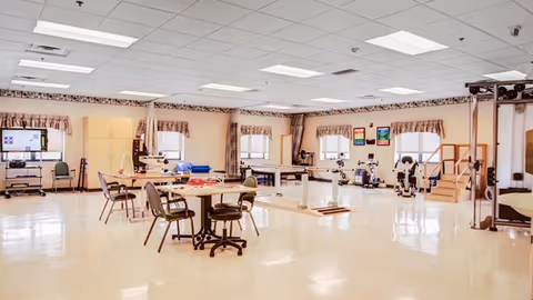 A spacious rehabilitation or therapy room with various exercise and therapy equipment, tables with chairs, and large windows with valance curtains allowing natural light to fill the room.