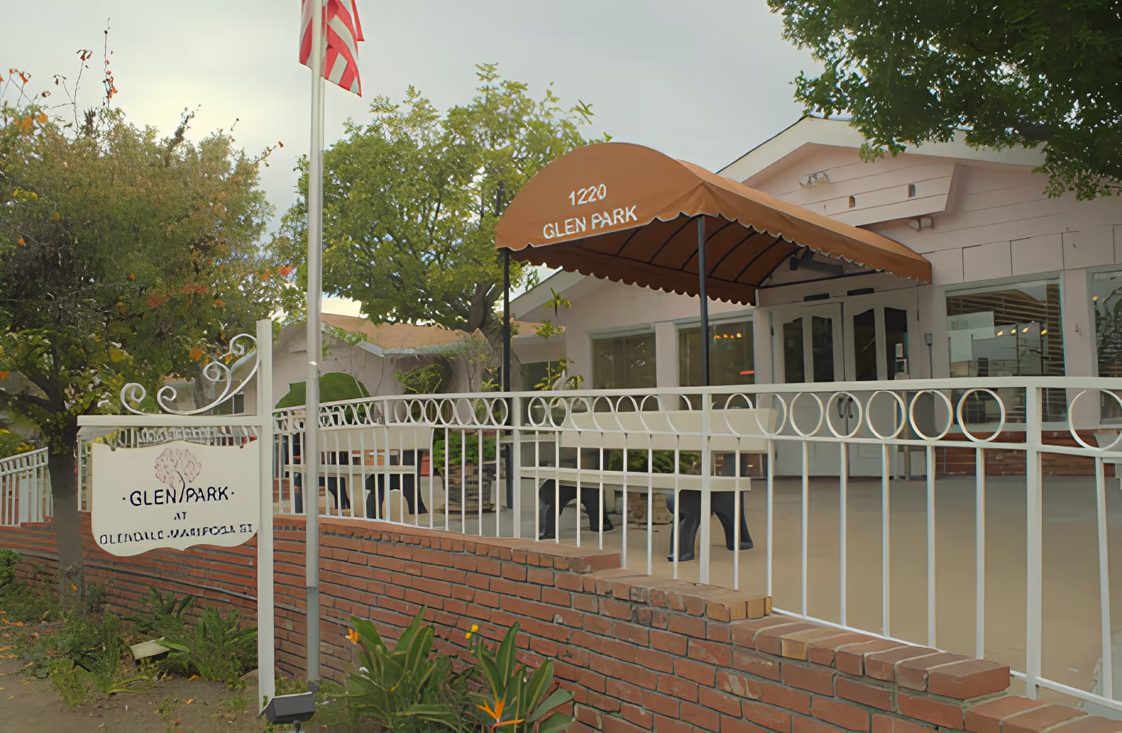 Front entrance of Glen Park senior living facility with a brown awning reading "1220 GLEN PARK", white fence and brick planter.