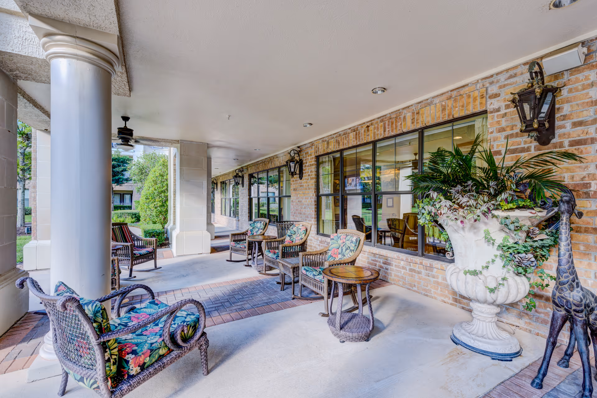 Covered outdoor patio area with wicker chairs featuring floral cushions, small round tables, large decorative planter with green plants, brick wall with windows, and ceiling fans.