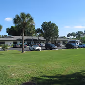 Exterior view of a single-story building with a parking lot in front, several cars parked, palm trees, and a large grassy area under a clear blue sky.