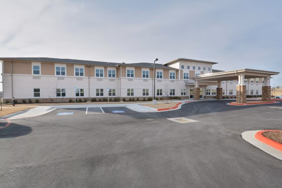 Exterior view of a two-story assisted living and memory care facility with a covered entrance supported by stone pillars. The building has multiple windows and a parking area with handicapped spaces in front.