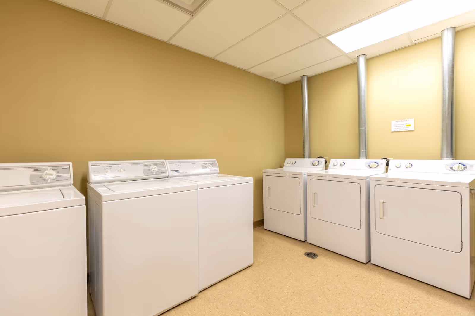 A clean laundry room with multiple white top-load washing machines and front-load dryers along beige walls.