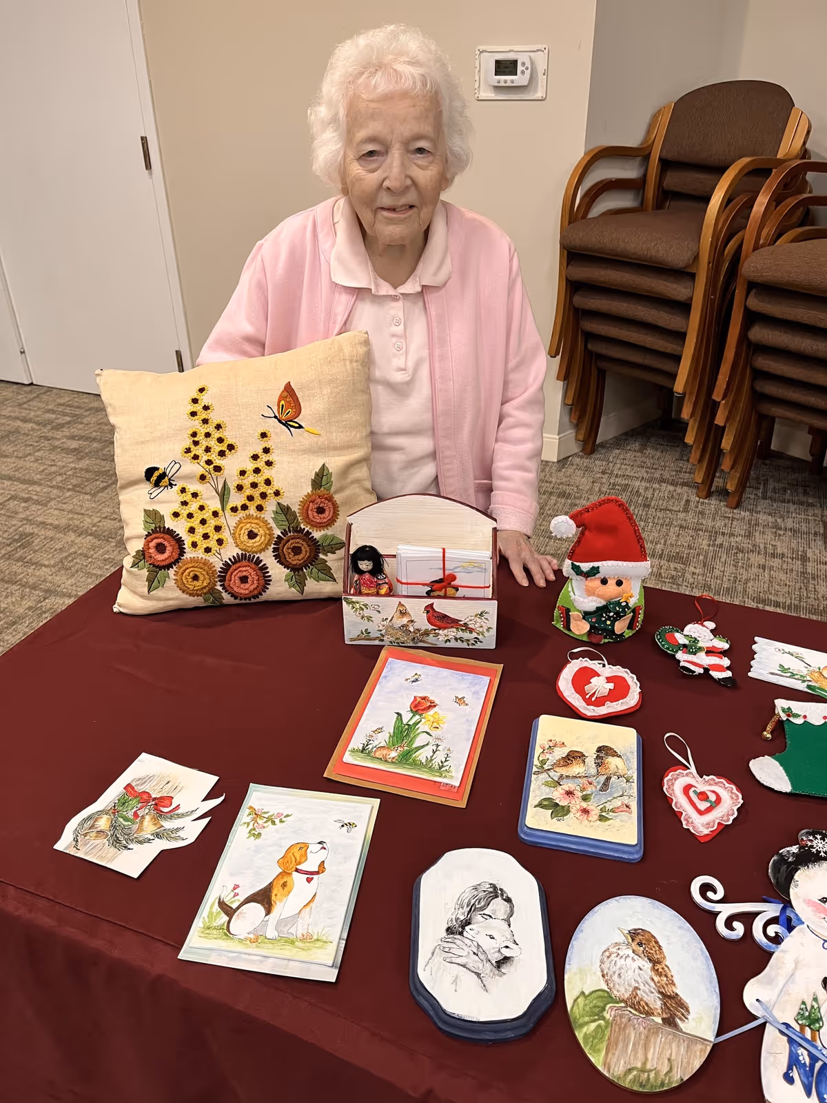 An elderly woman wearing a pink cardigan sits behind a table covered with a maroon cloth displaying various handmade crafts, including a floral embroidered pillow, painted cards, and Christmas-themed decorations. Behind her are stacked brown chairs and a beige wall with a thermostat.