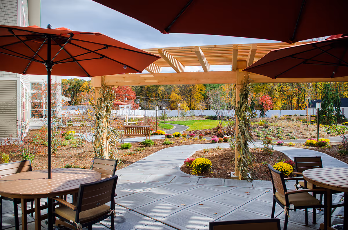 Outdoor patio area with round wooden tables and chairs under large red umbrellas. A wooden pergola decorated with dried corn stalks stands over a paved walkway that curves through a landscaped garden with colorful flowers and shrubs. Trees with autumn foliage are visible in the background.