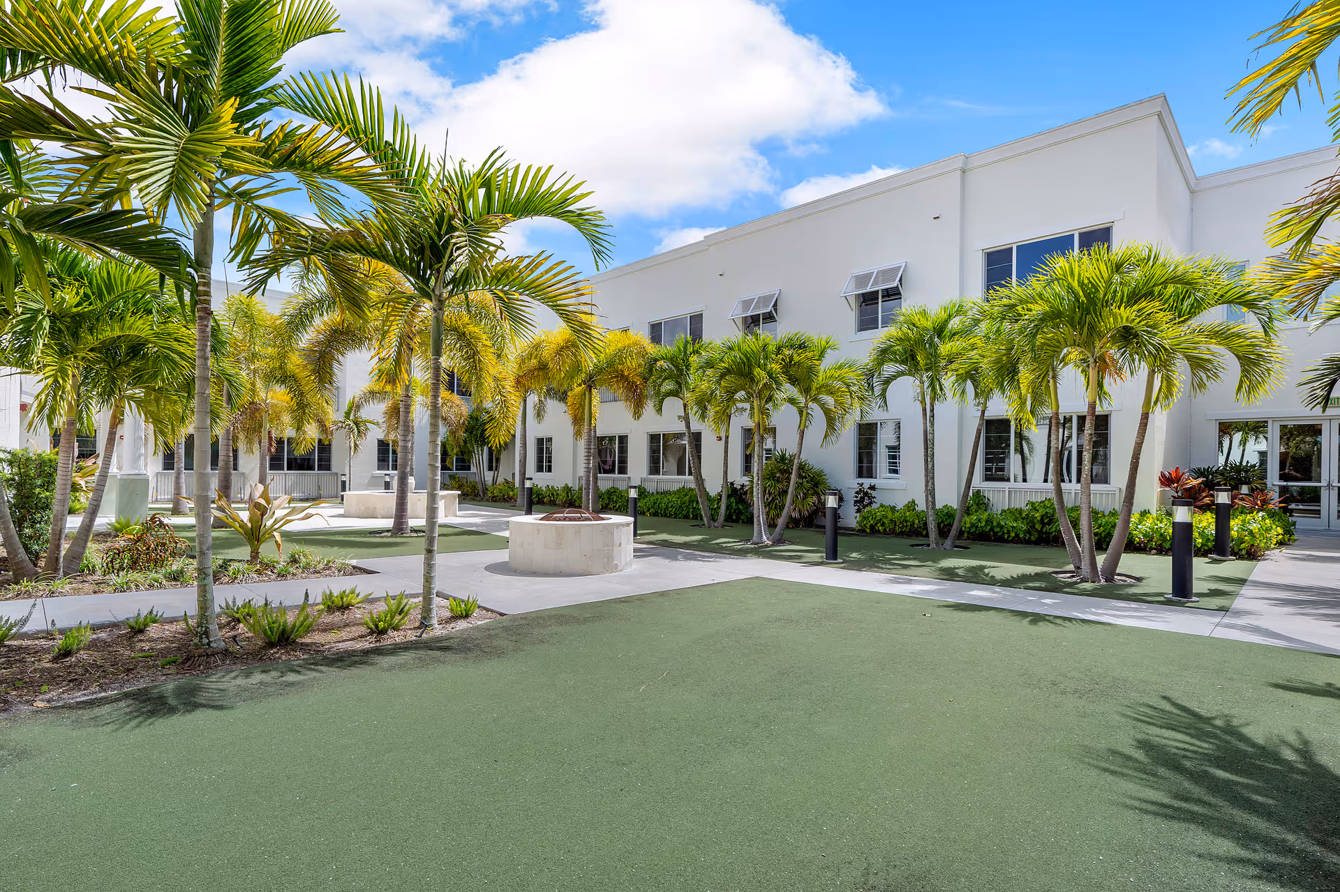 Sunny courtyard with palm trees, walkways, and seating in front of a white two-story building.