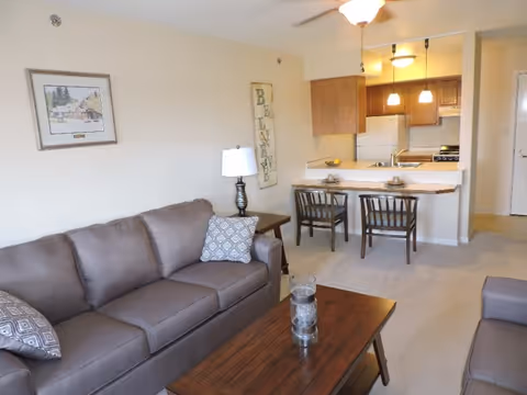 Furnished living room with sofas, a coffee table and lamp, opening to a kitchen with a breakfast bar and two chairs.