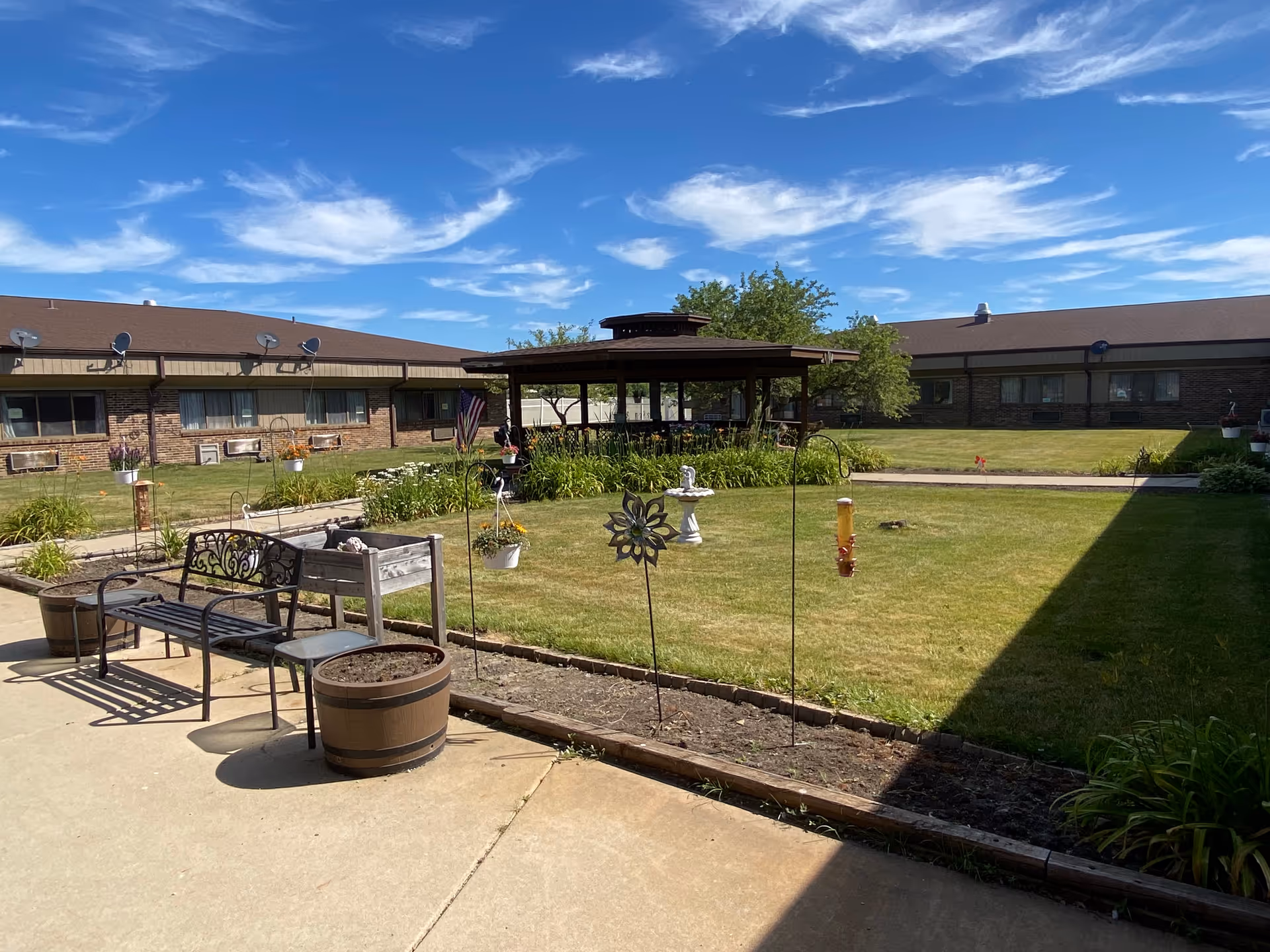 Outdoor courtyard area at Brickyard Healthcare - Merrillville Care Center featuring a grassy lawn with a central birdbath, a covered gazebo surrounded by plants, benches, potted plants, and hanging flower pots under a blue sky with scattered clouds.