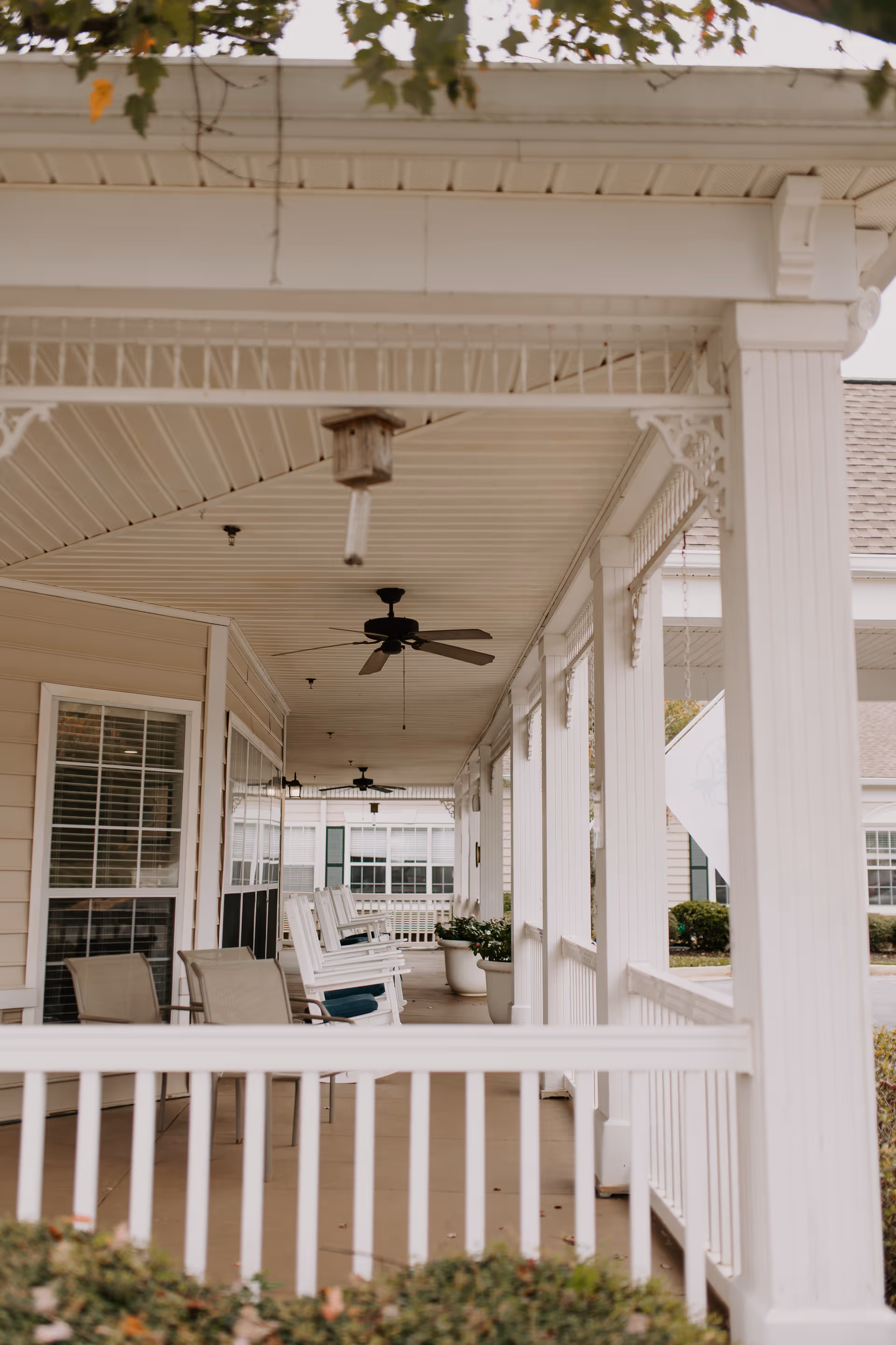 Covered porch area with white railings and columns, featuring several white rocking chairs with blue cushions and a couple of beige chairs. Ceiling fans and hanging birdhouses are visible on the porch ceiling. The porch overlooks a garden area with bushes and trees.