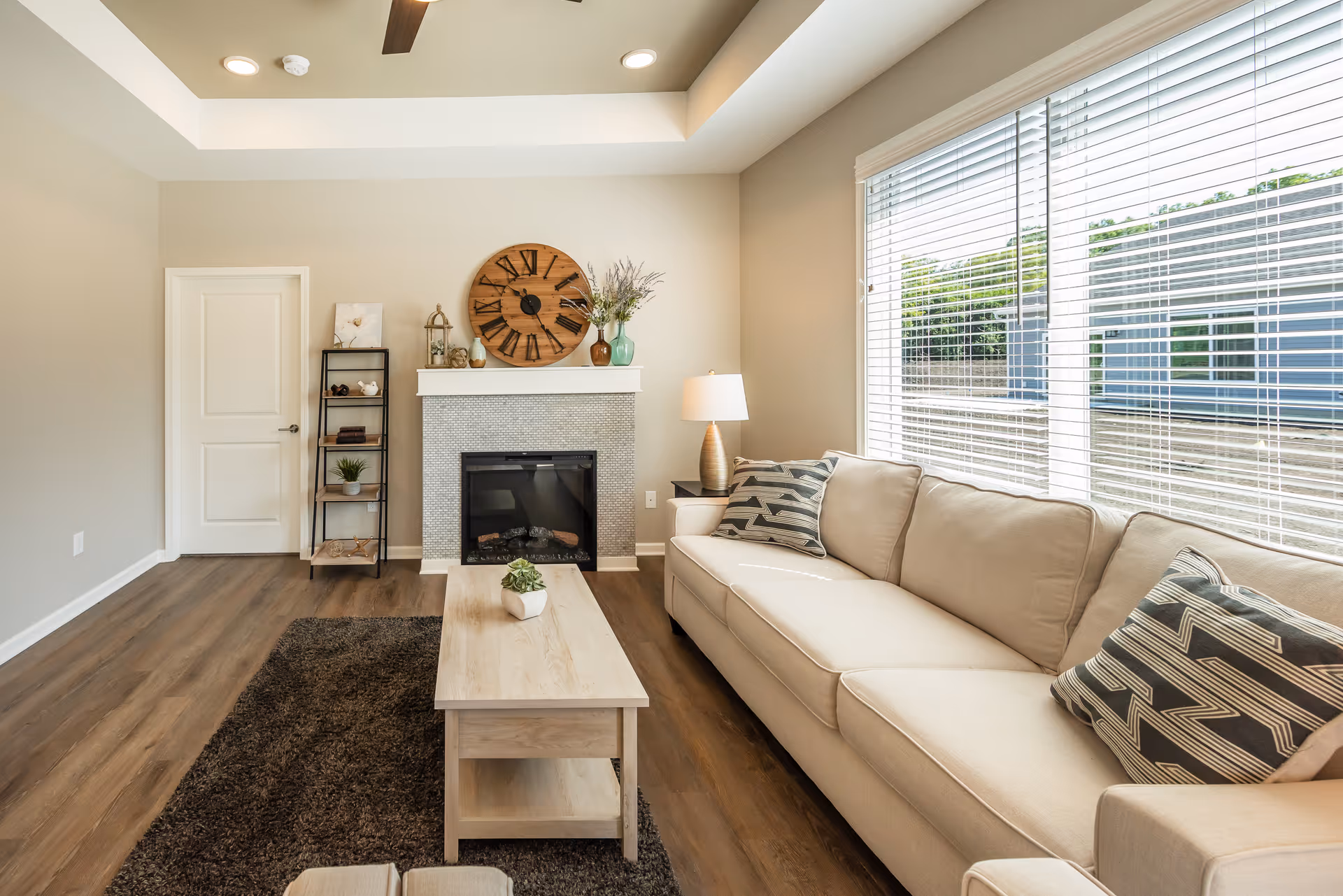 A bright living room with a beige sofa adorned with patterned pillows, a wooden coffee table with a small plant, a dark brown rug, and a fireplace with a large round wooden clock and decorative items on the mantel. A tall shelving unit stands next to a closed white door, and large windows with white blinds allow natural light to fill the room.
