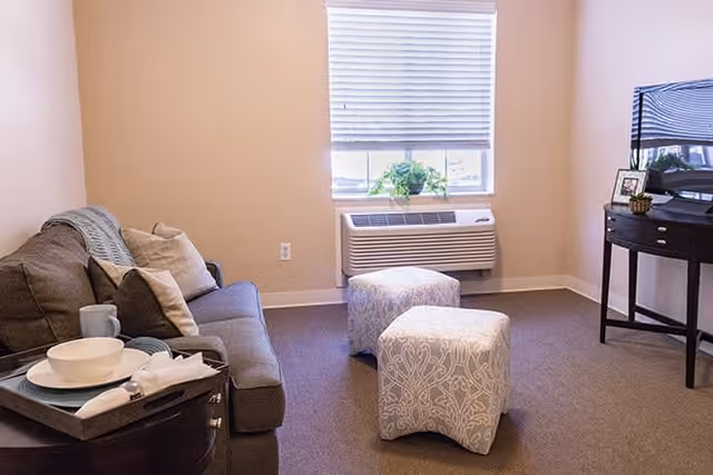 A cozy living room with a gray sofa adorned with pillows and a throw blanket, a small side table with a tray holding a bowl, cup, and napkin, two patterned ottomans in front of a window with blinds, and a flat-screen TV on a dark wooden stand.