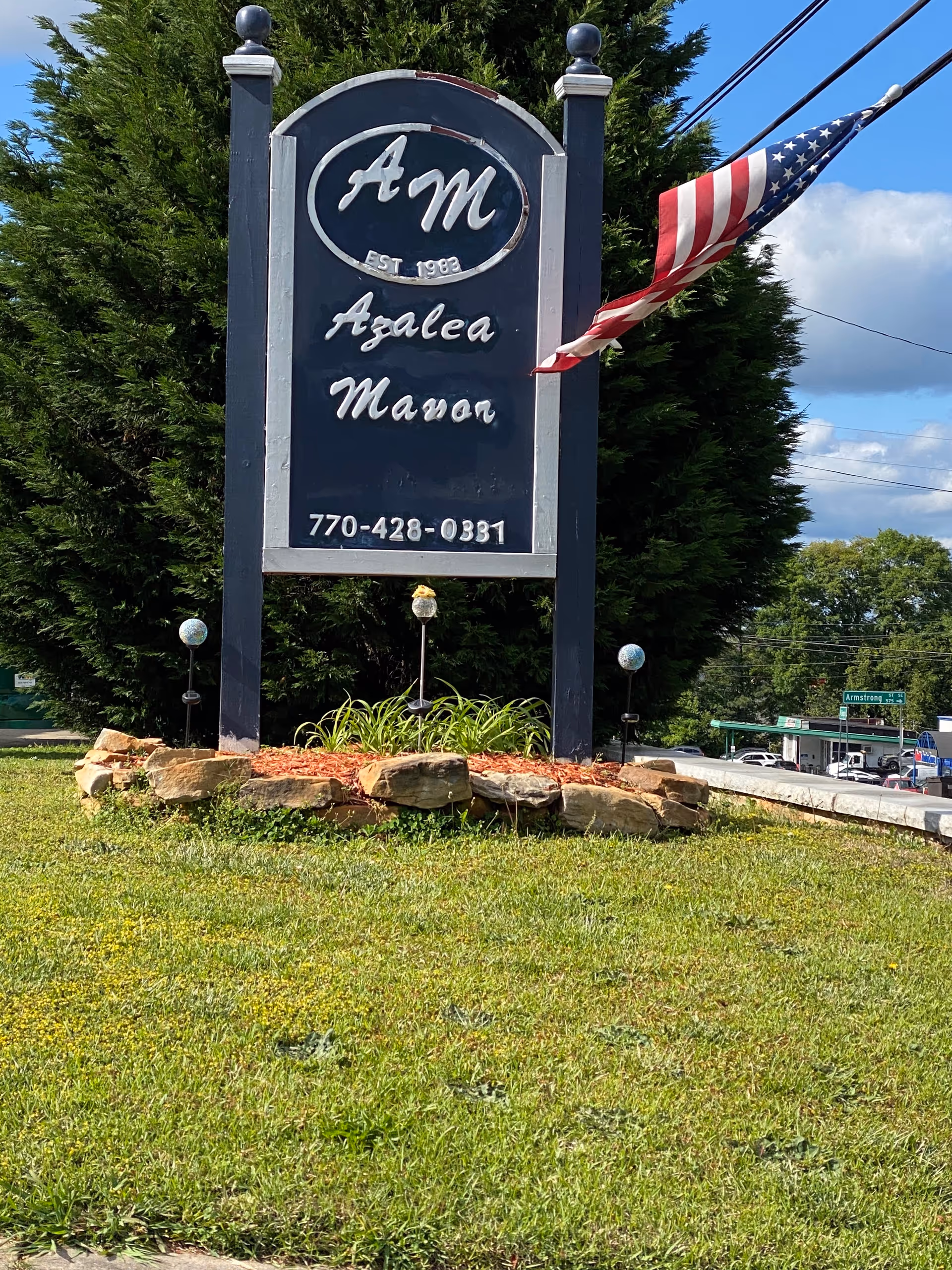 A large dark blue and white sign for Azalea Manor Senior Living with the initials 'AM' and the text 'Est. 1983' along with a phone number 770-428-0331. The sign is mounted on two posts and surrounded by a small landscaped area with rocks and plants. An American flag is attached to the right post, waving in the wind. There are trees and a street with cars in the background under a partly cloudy sky.