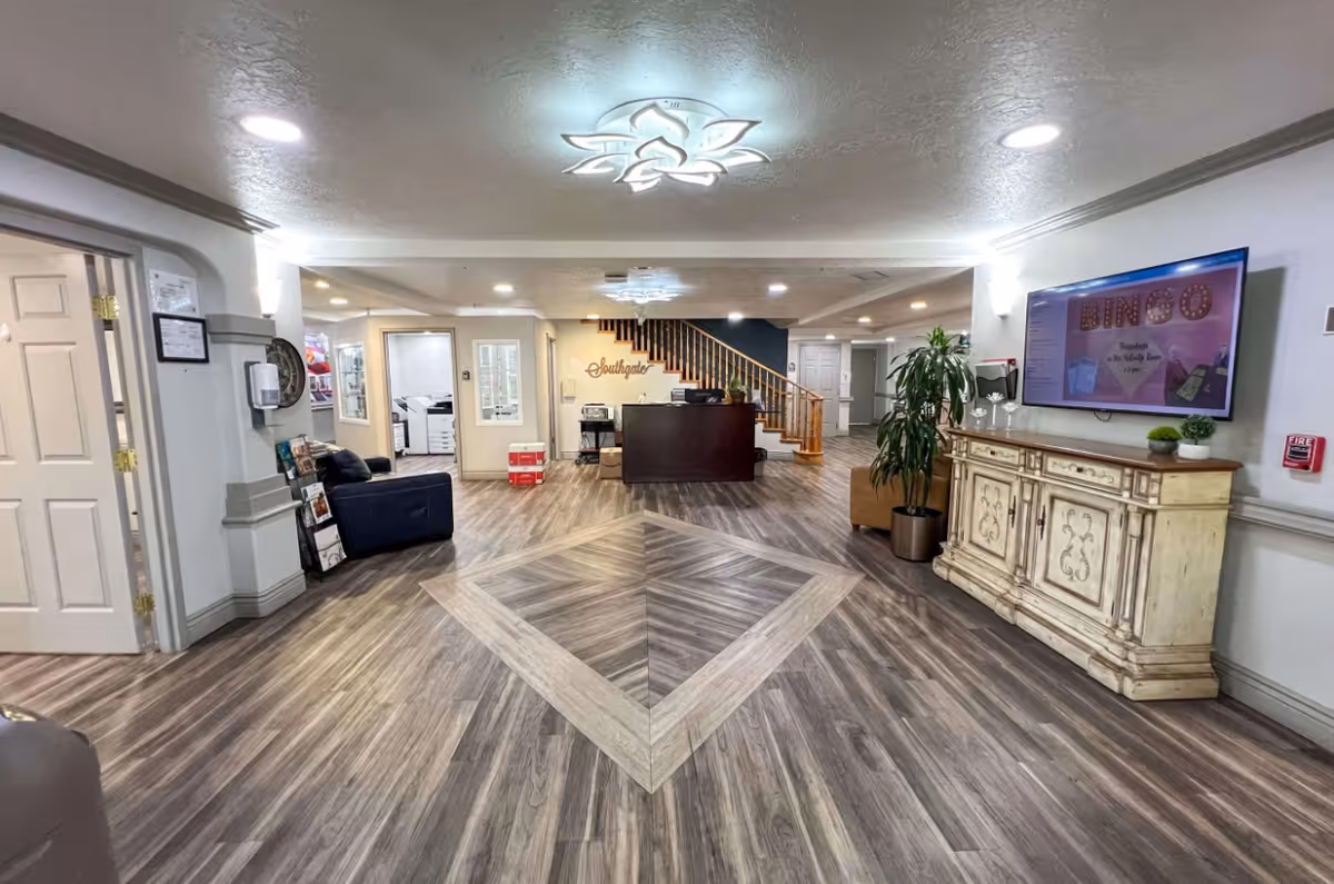 Interior view of a senior living facility lobby with wood-patterned flooring, a reception desk in the center, a staircase behind it, a decorative ceiling light, a TV screen on the right wall displaying a bingo announcement, a vintage-style cabinet below the TV, and seating areas on the left side.
