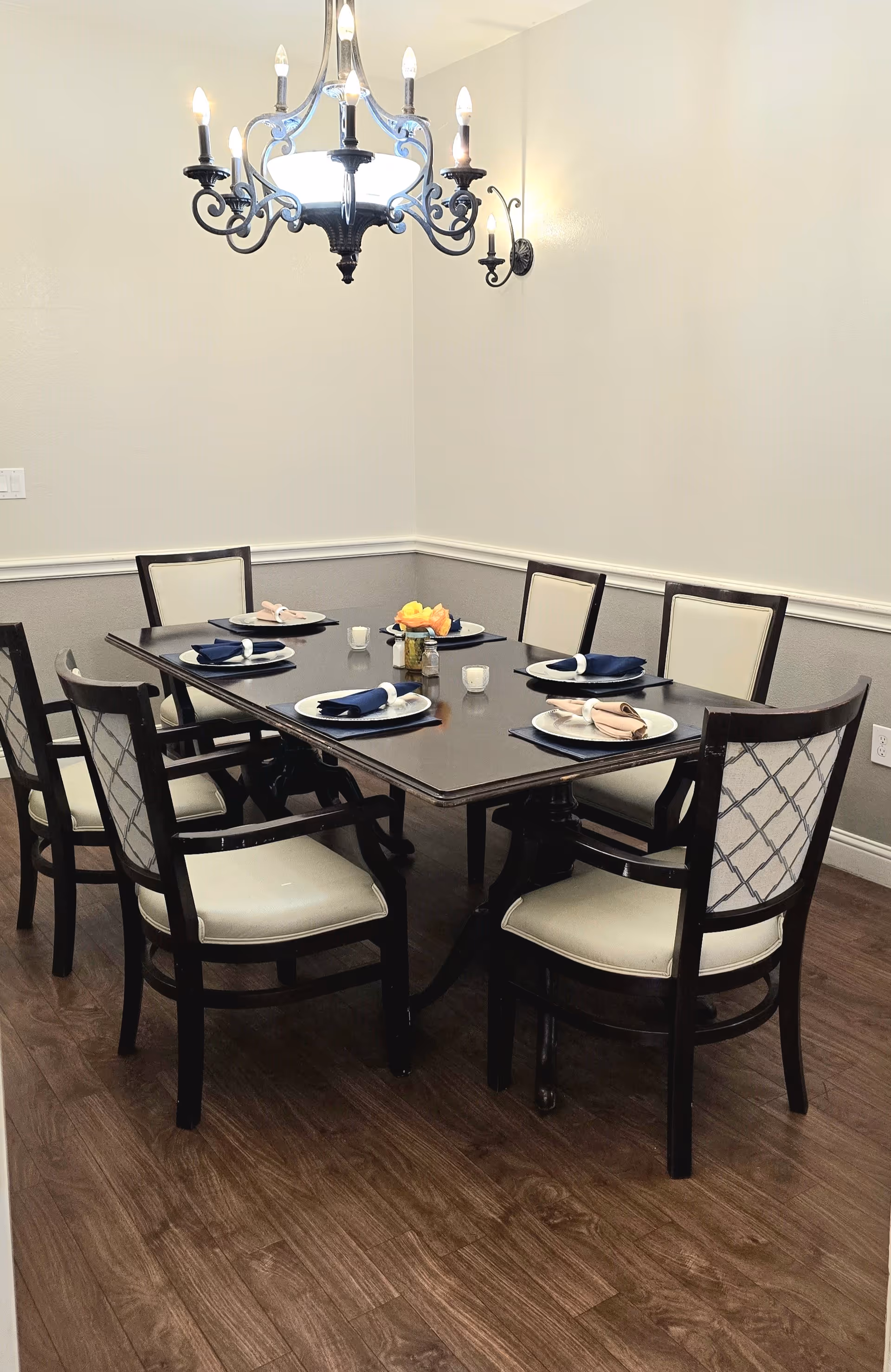 A dining room with a dark wooden rectangular table set for six people. Each place setting has a white plate, a folded napkin in either navy blue or beige, and a small candle. The chairs have dark wooden frames with cream-colored cushions and patterned backs. A decorative chandelier with multiple lights hangs above the table, and the room has light-colored walls with a chair rail and wood flooring.