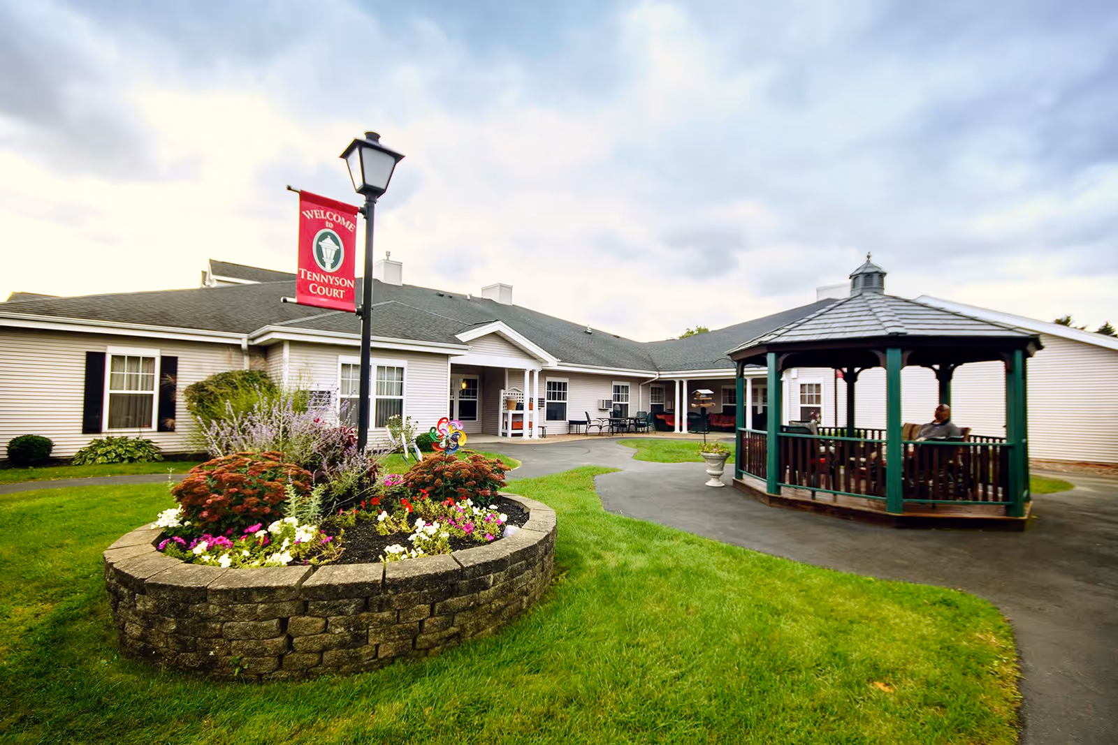Outdoor courtyard area of Tennyson Court featuring a circular flower bed with colorful flowers and a lamppost with a red banner that reads 'Welcome to Tennyson Court.' There is a paved walkway surrounding the flower bed and leading to a gazebo with green pillars and a gray roof, where a person is seated. The building in the background is a single-story structure with white siding and multiple windows.