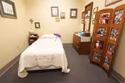 A small, cozy room with a single bed covered with a white bedspread. The walls are decorated with framed pictures and artwork. There is a wooden dresser with various items on top and a decorative folding screen with floral patterns. The room has a carpeted floor and soft lighting.