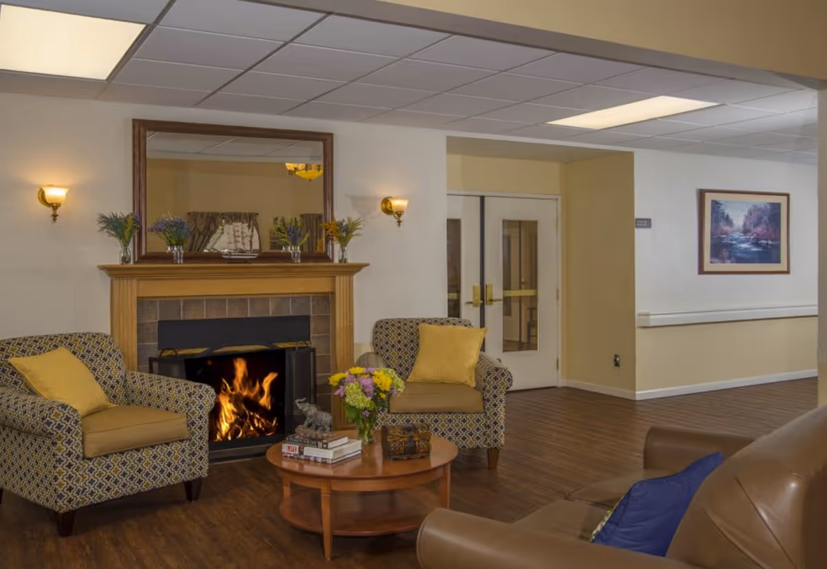 A cozy senior living facility common area with two patterned armchairs with yellow cushions, a wooden coffee table with books, flowers, and a small elephant figurine, a lit fireplace with a wooden mantle decorated with vases of flowers and a model ship, a large mirror above the fireplace, and a leather sofa with a blue cushion. The room has wood flooring, beige walls, wall sconces, and double doors in the background.