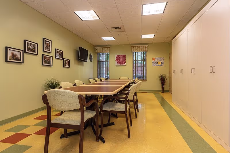 A well-lit room with a long rectangular table surrounded by cushioned chairs. The walls are painted light green and decorated with framed pictures and two windows with valances. There is a flat-screen TV mounted on the wall and a row of white cabinets along one side of the room. The floor has a colorful pattern with yellow, green, and red tiles.